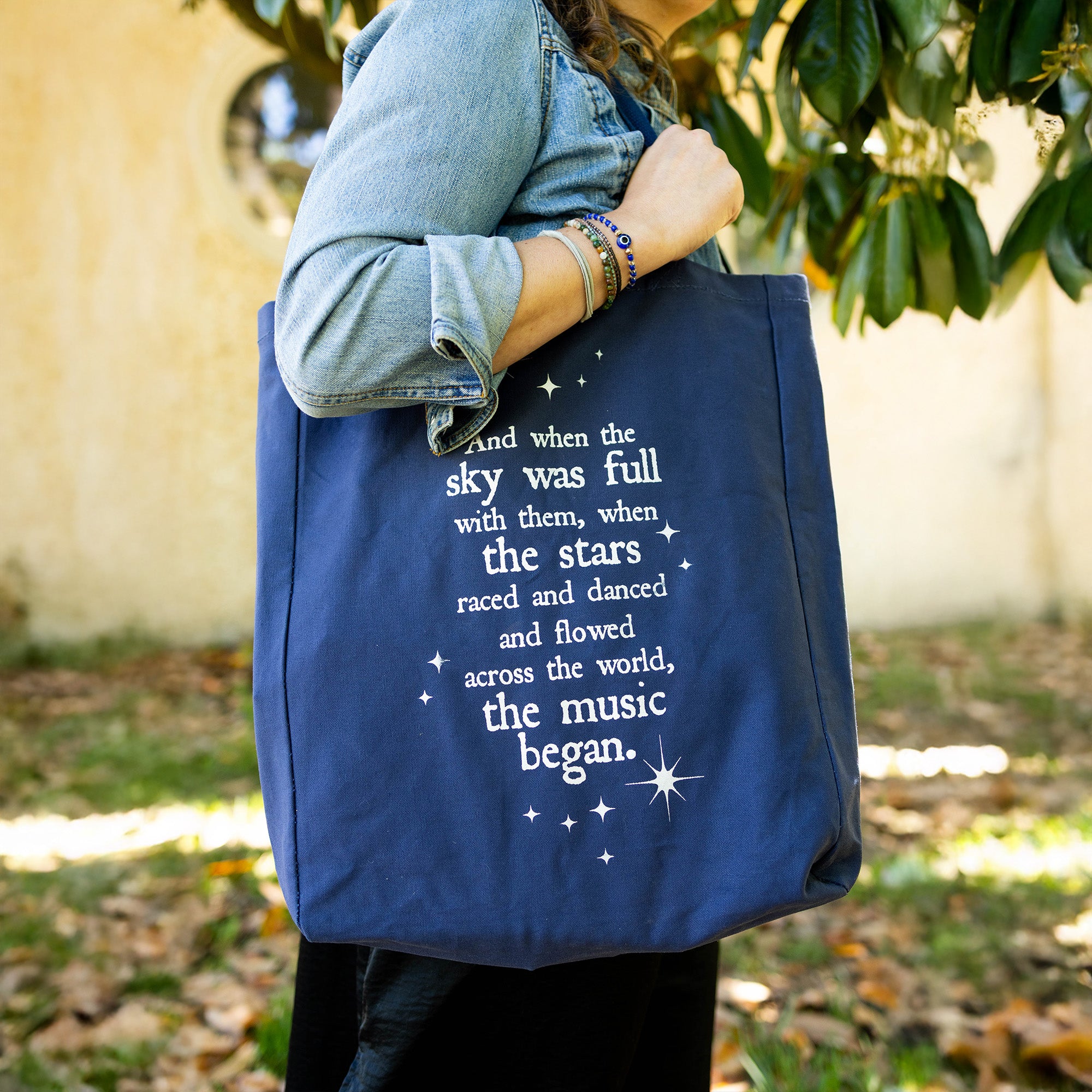 model carrying navy tote that says 'AND WHEN THE SKY WAS FULL WITH THEM, WHEN THE STARS RACES AND DANCED AND FLOWERS ACROSS THE WORLD, THE MUSIC BEGAN'
