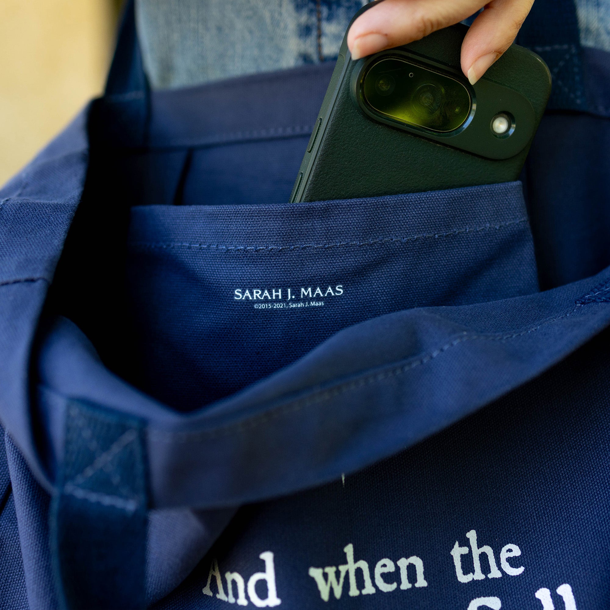 Person holding a phone over a blue tote with 'Sarah J. Maas' branding.