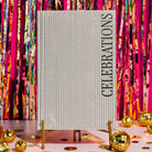 A vertical hardcover notebook with a cream-colored cover featuring thin vertical tan stripes and the word 'CELEBRATIONS' printed in black serif font along the right side, shown on a plain white background.