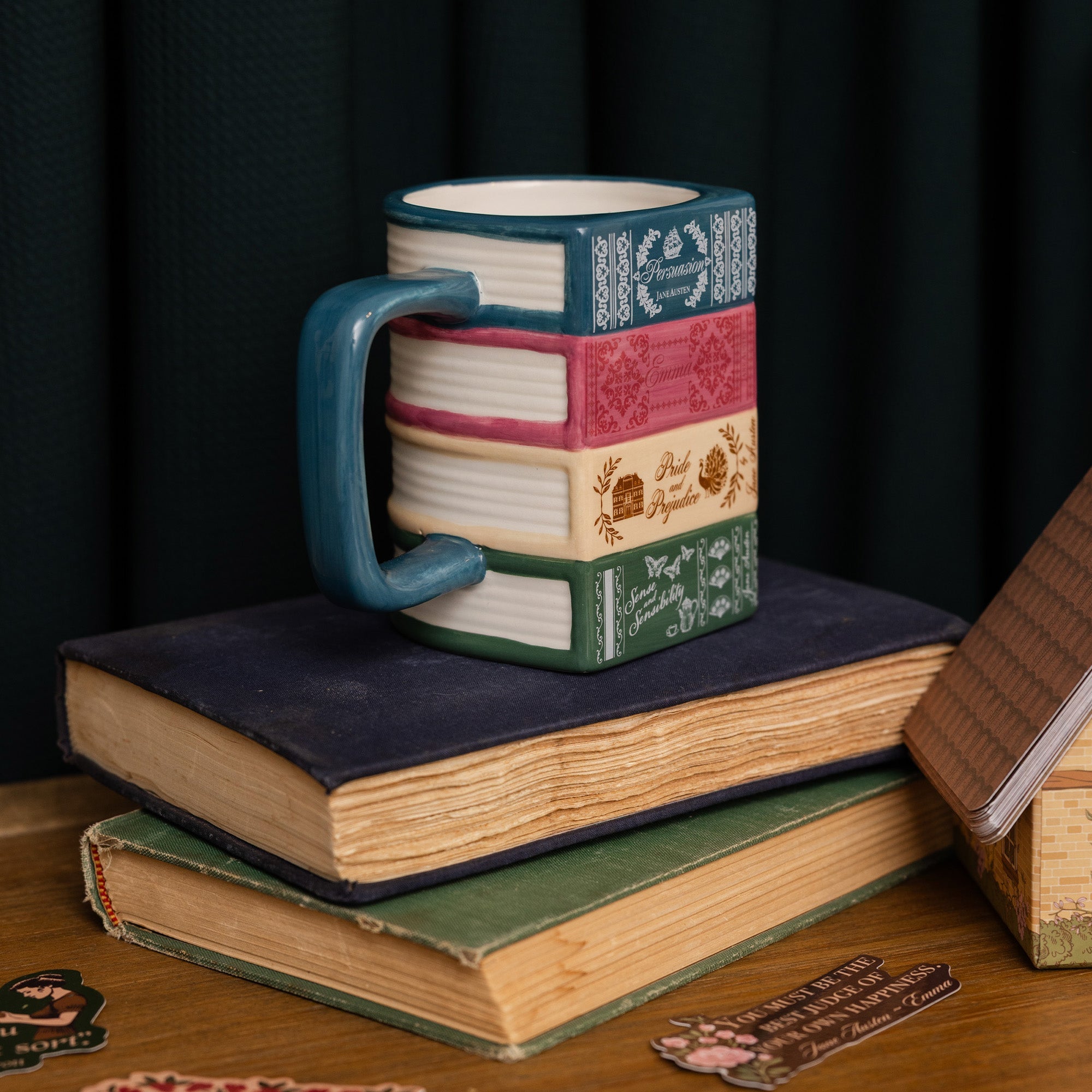 A ceramic mug shaped like a stack of vintage books with a teal handle, displayed on top of stacked antique books against a dark curtain background.