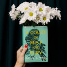 model holding 'A Court of Mist and Fury' by Sarah J. Maas vase with a green cover and white spine and white flowers inside