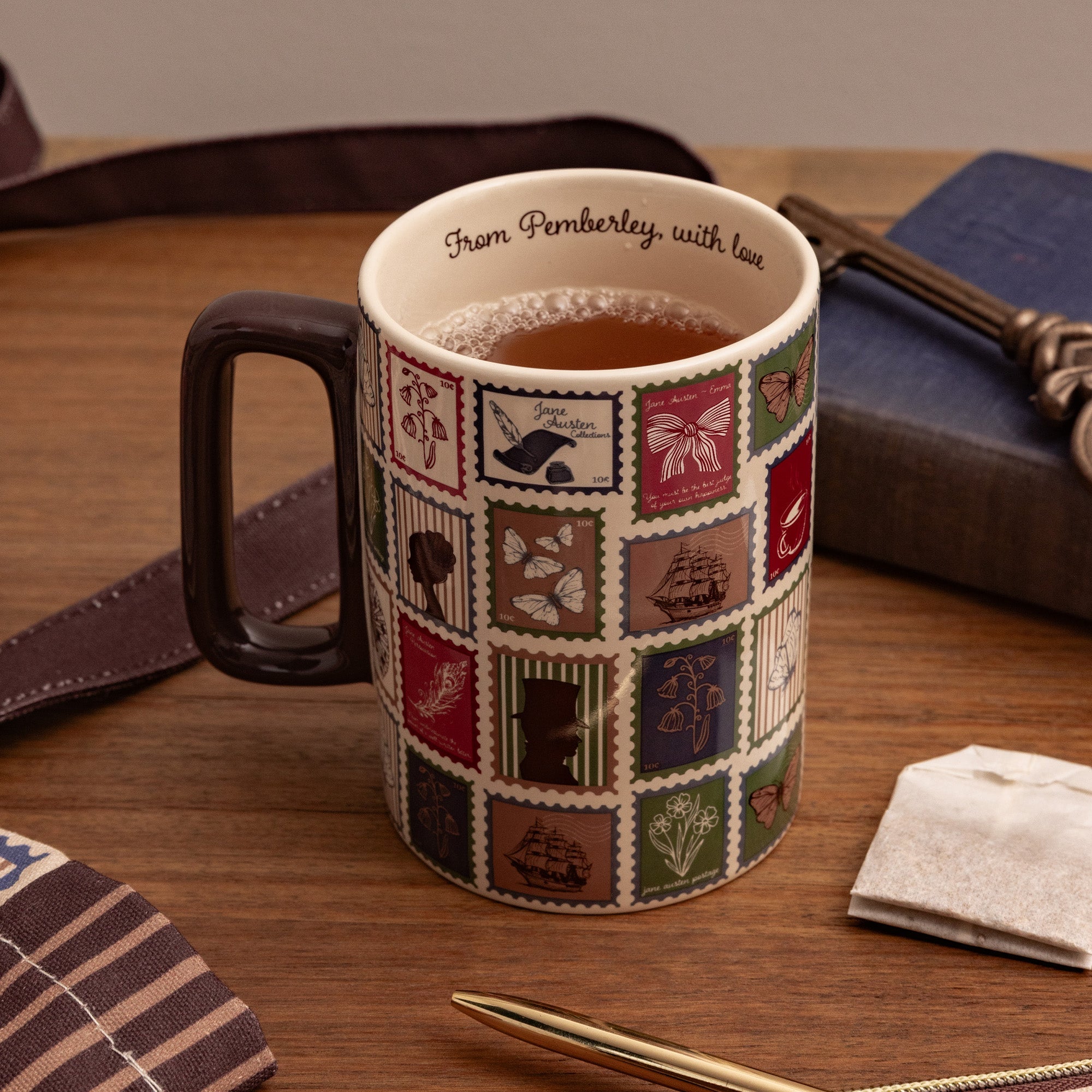 A ceramic mug filled with tea sits on a wooden table, decorated with a patchwork of vintage postage stamp designs in muted red, green, blue, and brown tones. The mug has a dark brown handle and the inside rim is printed with the text 'From Pembrory, with love'.