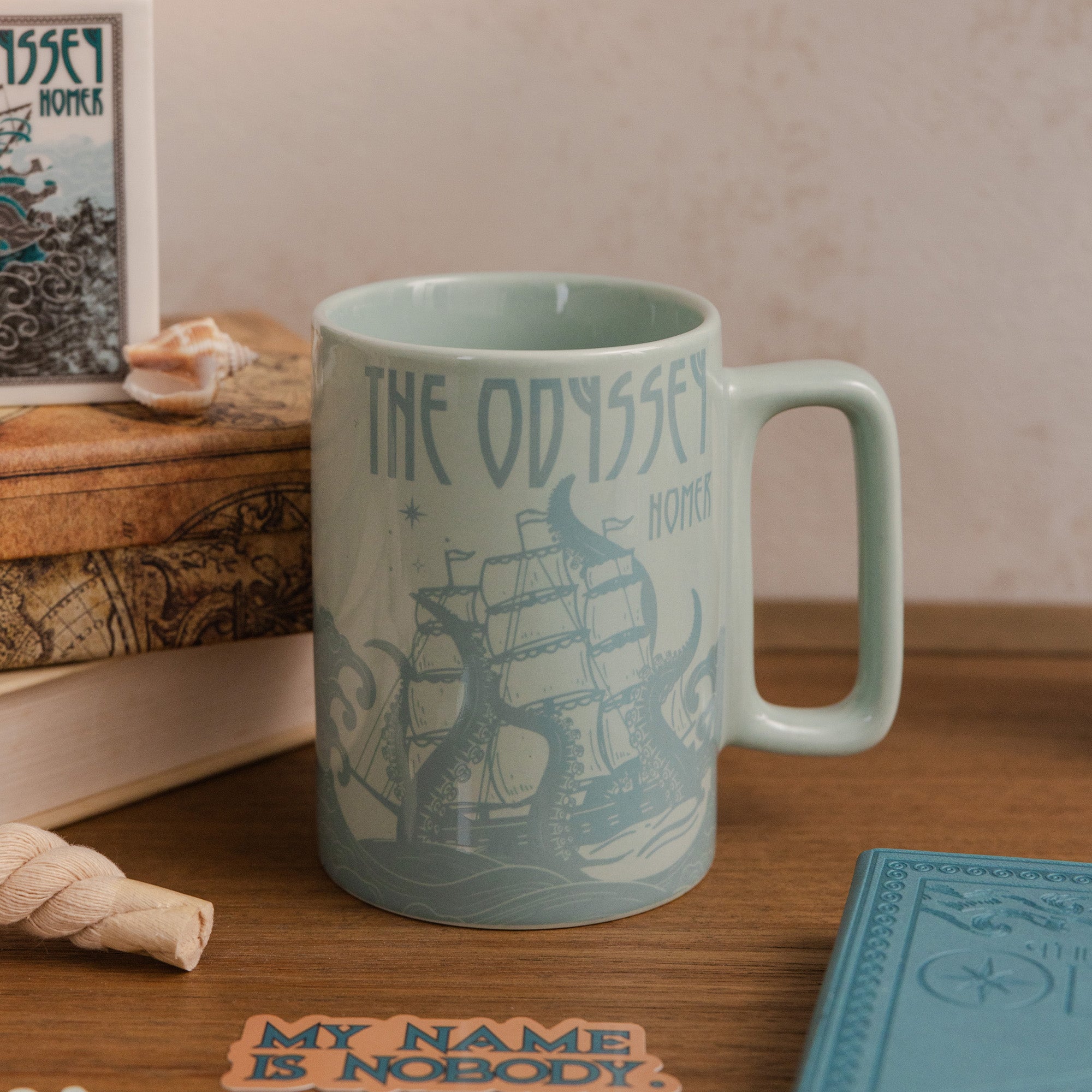 Mug with 'The Odyssey' design on a wooden surface with books and a shell.