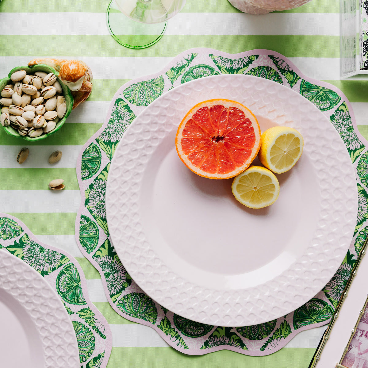 A styled tabletop setting with a pink textured plate on top of a green and white patterned placemat, set on a green and white striped tablecloth. Slices of grapefruit, lemon, and pistachios are arranged on the plate, with additional green glassware and food props around it.