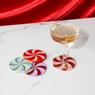 Four round spiral-patterned coasters in red, pink, blue, and purple are arranged on a white marble surface next to a coupe glass filled with a pale amber drink, with a draped red fabric backdrop.