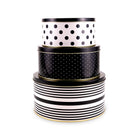 Three stacked round gift tins with black and white patterns, featuring polka dots and stripes, with thin gold trim, on a white background.