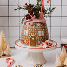 A gingerbread house-shaped ceramic jar or container with a pink lid, decorated with white snowflakes, candy canes, and a brown door, displayed on a white pedestal stand with peppermint candies and small decorative trees.