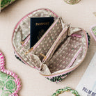 A zippered travel pouch with a geometric patterned fabric lining and a black patterned exterior, shown open with a passport inside. The pouch is photographed on a light beige textured surface with other travel accessories partially visible around it.