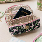 A patterned zippered pouch with pink trim and a gold logo emblem, shown open with a passport inside on a beige fabric surface.