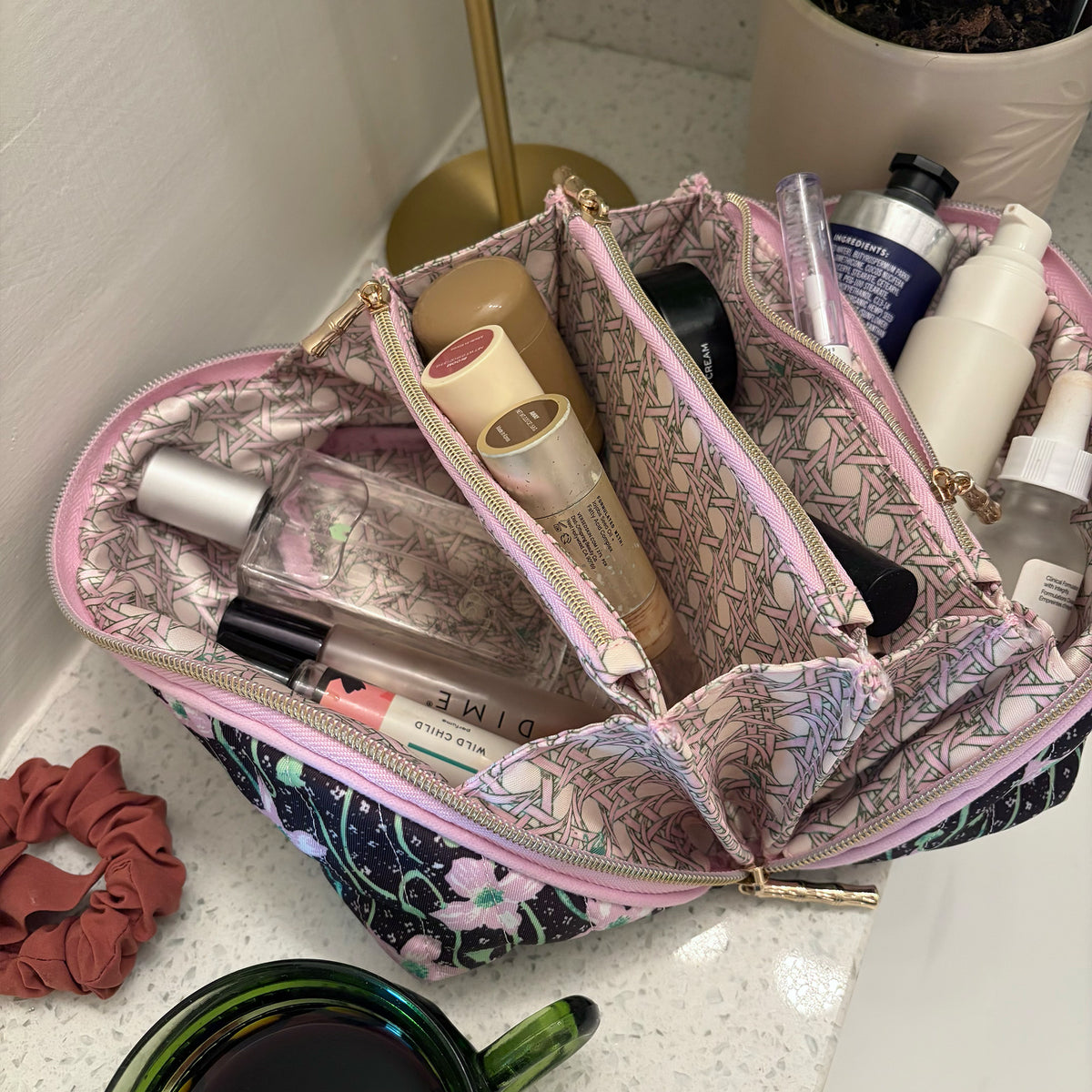 A patterned zippered pouch with pink trim and a gold logo emblem, shown open with a passport inside on a beige fabric surface with makeup inside