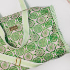 A close-up of a tote bag featuring a green and white floral pattern on a light pink background, with a light green and white striped handle and strap, and a gold-tone brand plaque.