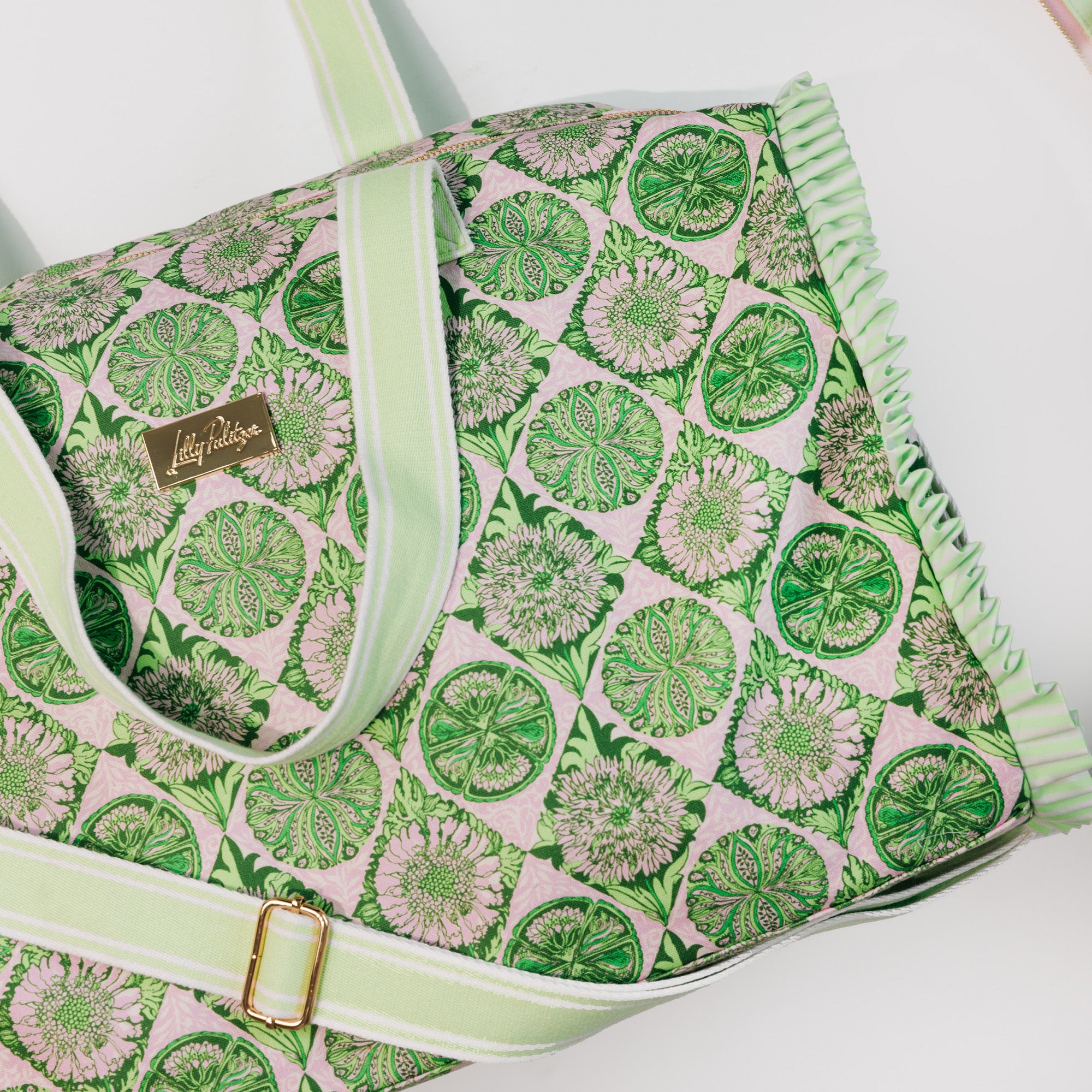 A close-up of a tote bag featuring a green and white floral pattern on a light pink background, with a light green and white striped handle and strap, and a gold-tone brand plaque.