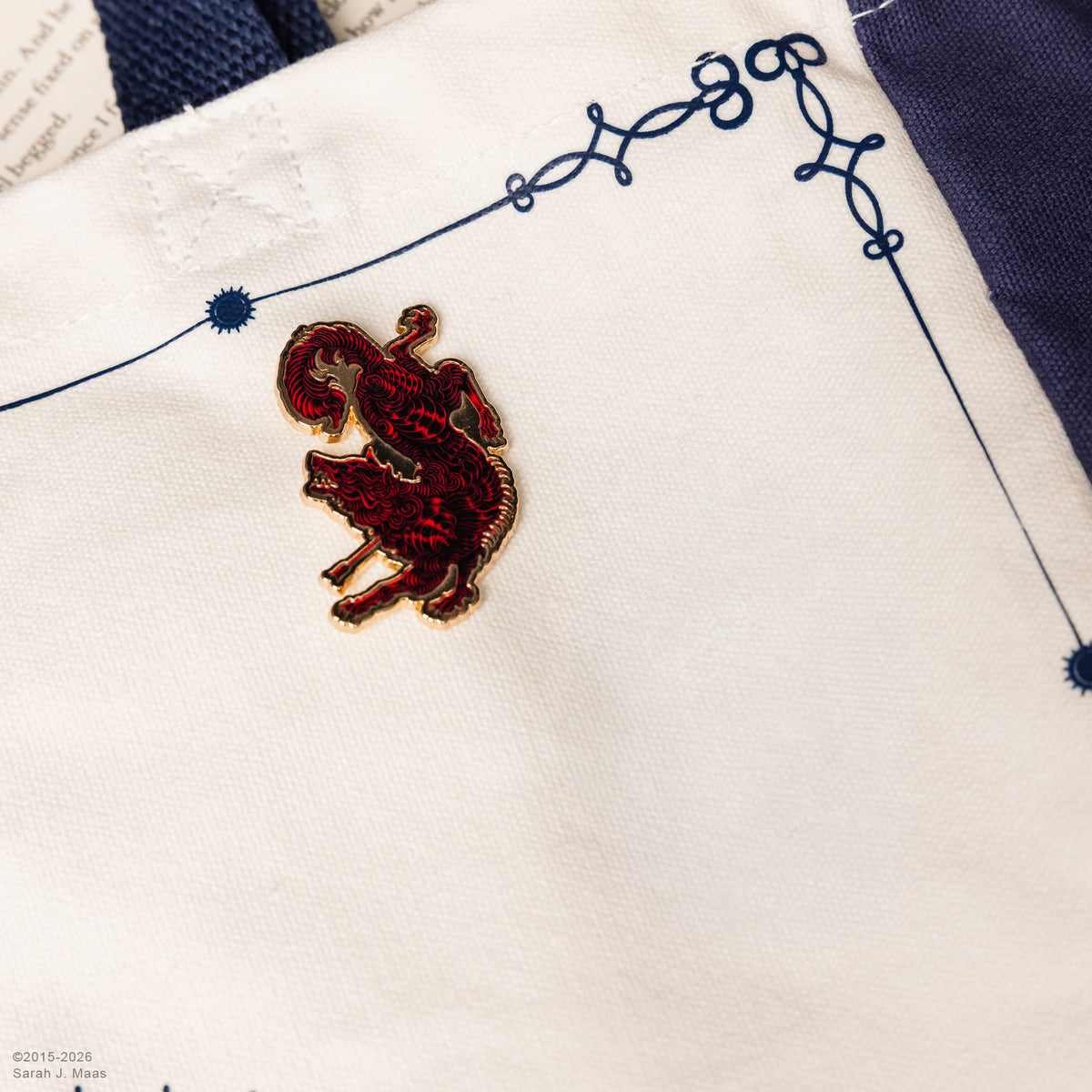 A gold-plated ACOTAR enamel lapel pin depicting a stylized red and black creature on a tote bag