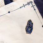 A hand enamel pin with a gold-tone metal border and a dark blue background decorated with gold celestial patterns, stars, and a central eye motif on a tote bag