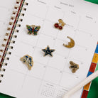 Kate Spade New York enamel pin set in a green presentation box, featuring six pins: cherries, a star, a bee, a crescent moon, a butterfly, and a bird on a planner