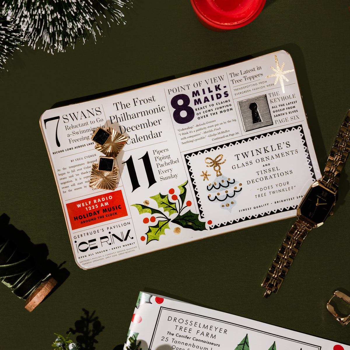 A flat-lay of a vintage-style Christmas  tray featuring retro newspaper clippings, holiday illustrations, and gold and black pins, styled with a gold watch, pinecones, and a red wax seal on a dark olive green background.