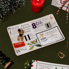 side view of a vintage-style Christmas  tray featuring retro newspaper clippings, holiday illustrations, and gold and black pins, styled with a gold watch, pinecones, and a red wax seal on a dark olive green background.