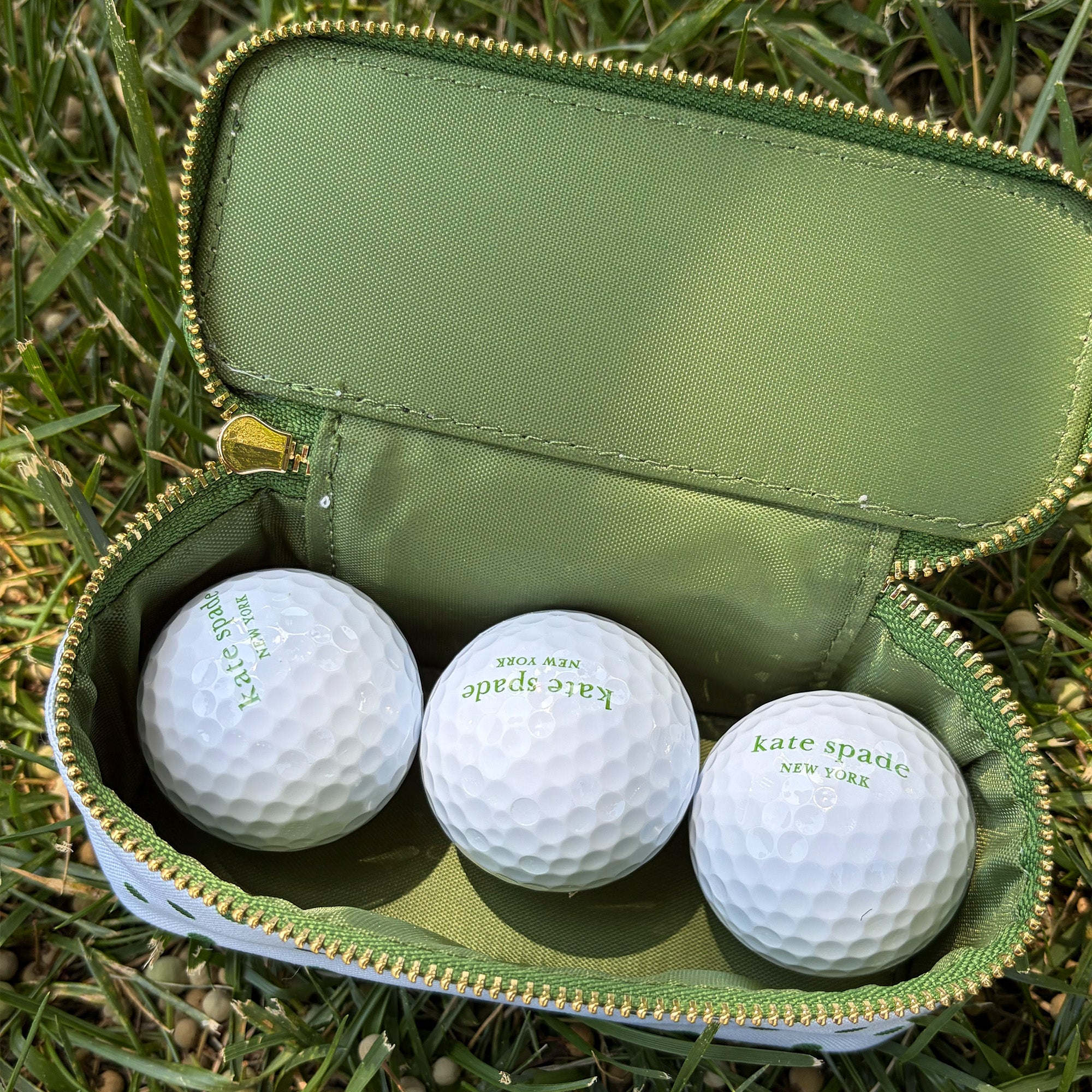 Three white golf balls with green 'kate spade NEW YORK' logos inside an open olive green zippered carrying case with gold zipper trim, placed on green grass.