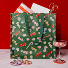 A green reusable tote bag with a candy print pattern, styled on a white marble surface with a red background. A pair of gold scissors is tucked into the top opening, with a striped paper insert and a glass coupe of peppermint candy nearby.