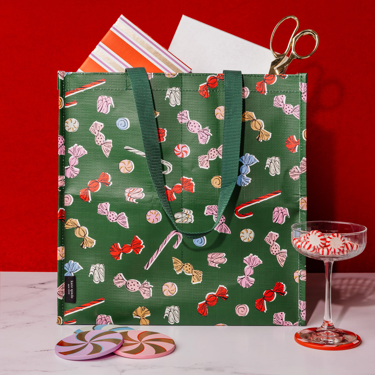 A green reusable tote bag with a candy print pattern, styled on a white marble surface with a red background. A pair of gold scissors is tucked into the top opening, with a striped paper insert and a glass coupe of peppermint candy nearby.