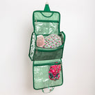 A green and white striped hanging toiletry organizer with multiple zippered compartments with items inside on a grey background