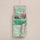 A hanging pink and green striped travel hairbrush organizer pouch with clear pockets containing hairbrushes, shown against a neutral textured beige background.