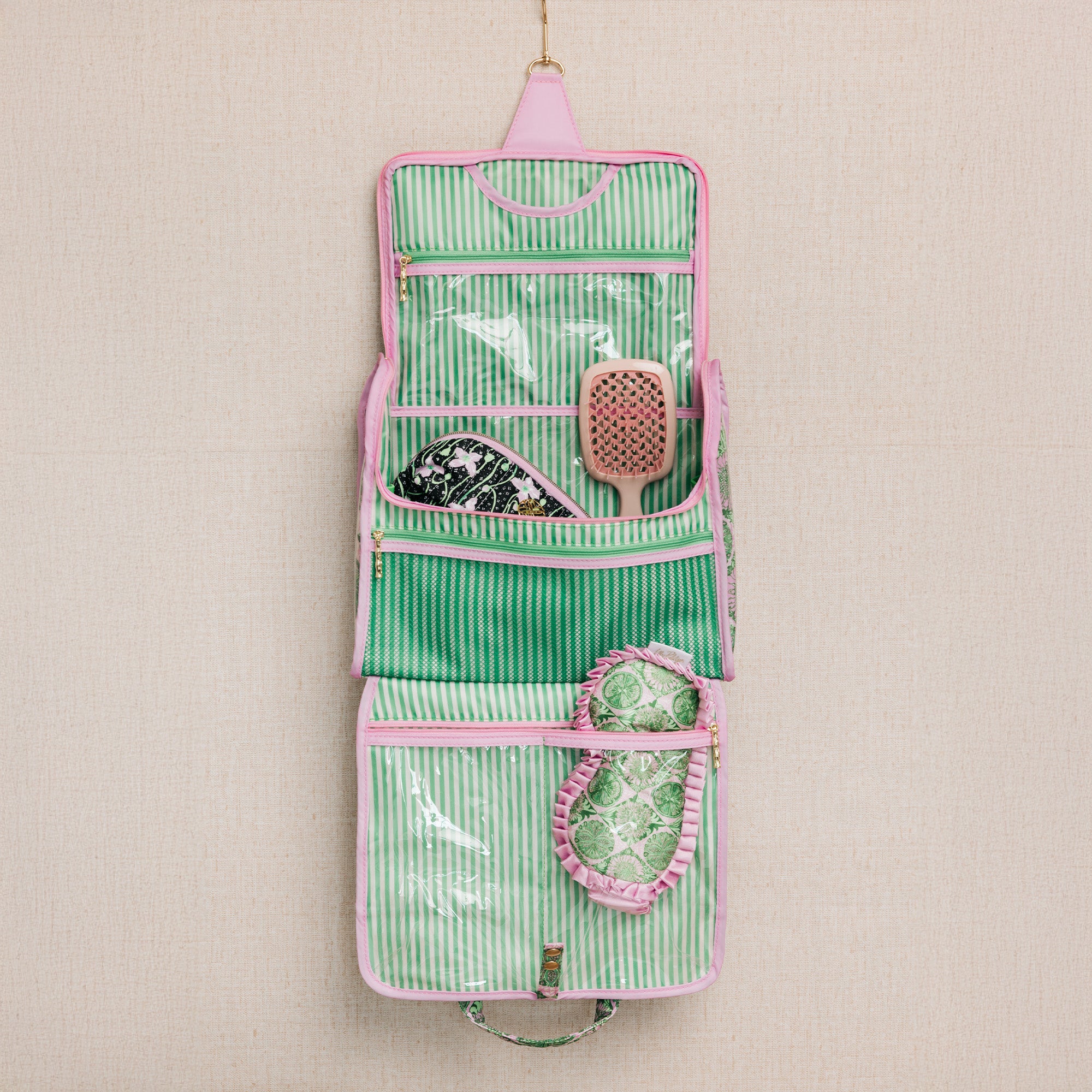 A hanging pink and green striped travel hairbrush organizer pouch with clear pockets containing hairbrushes, shown against a neutral textured beige background.