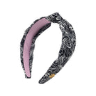 A twisted fabric headband featuring a black and white zebra print on the exterior and a solid light pink interior, with a small gold logo charm.