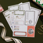 A fan of printed holiday invitation cards or stationery sheets laid on a dark green surface, surrounded by ribbon and gift props. The cards feature festive borders, gold script text, and holiday-themed designs including a Christmas tree and a coral 'Happy Holidays' box.