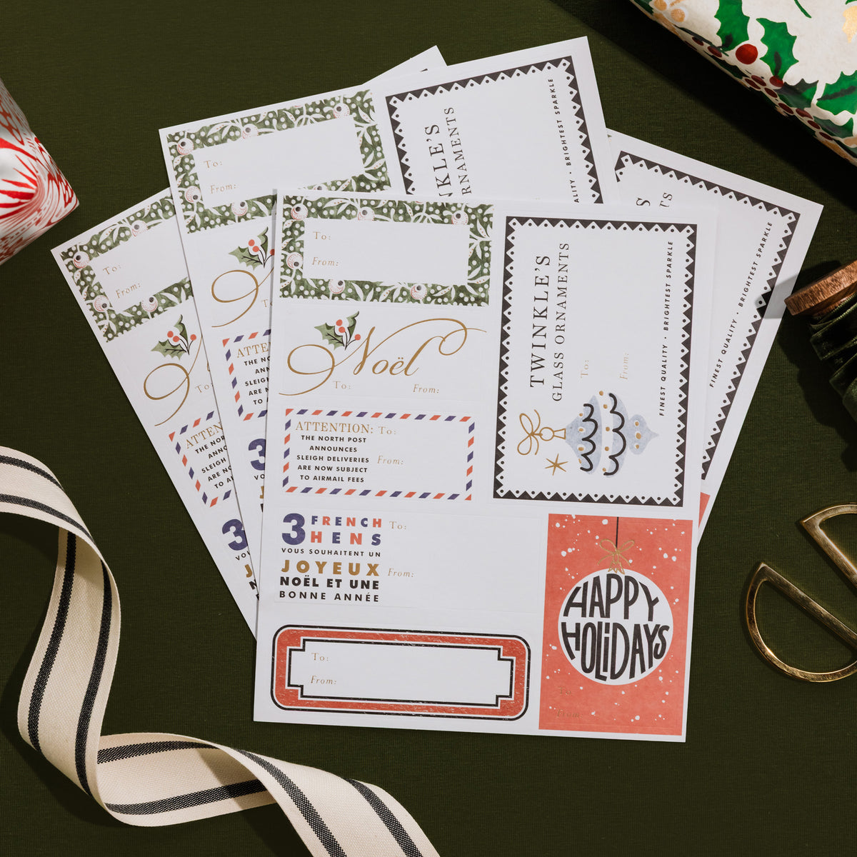 A fan of printed holiday invitation cards or stationery sheets laid on a dark green surface, surrounded by ribbon and gift props. The cards feature festive borders, gold script text, and holiday-themed designs including a Christmas tree and a coral 'Happy Holidays' box.