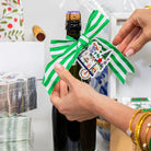 Person wrapping a bottle with a green and white striped ribbon and decorative label.