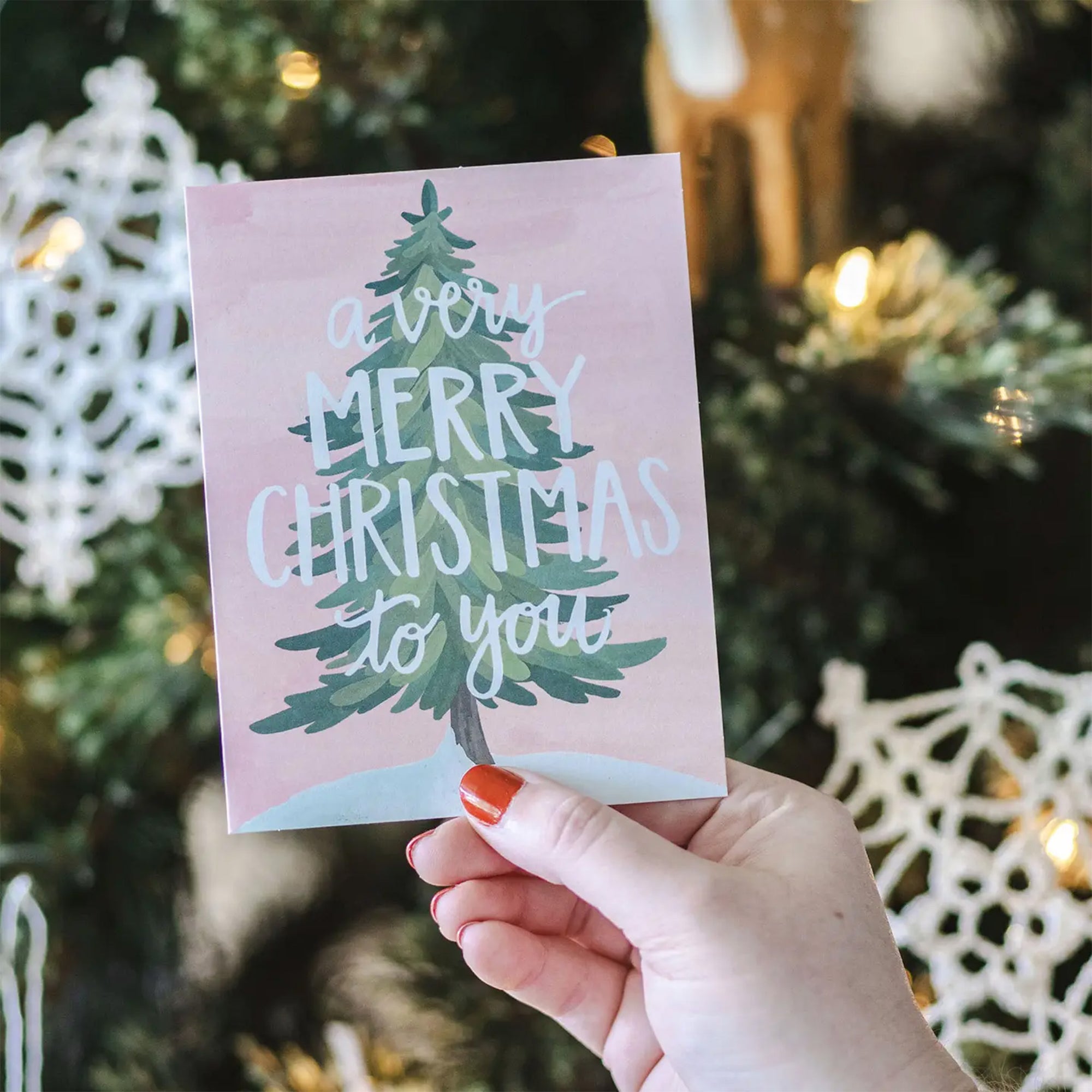 Hand holding a Christmas card with a tree and 'Merry Christmas' text in front of a decorated Christmas tree.