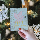 Hand holding a Christmas card with a dove and 'Peace to you and yours' text in front of a decorated tree.