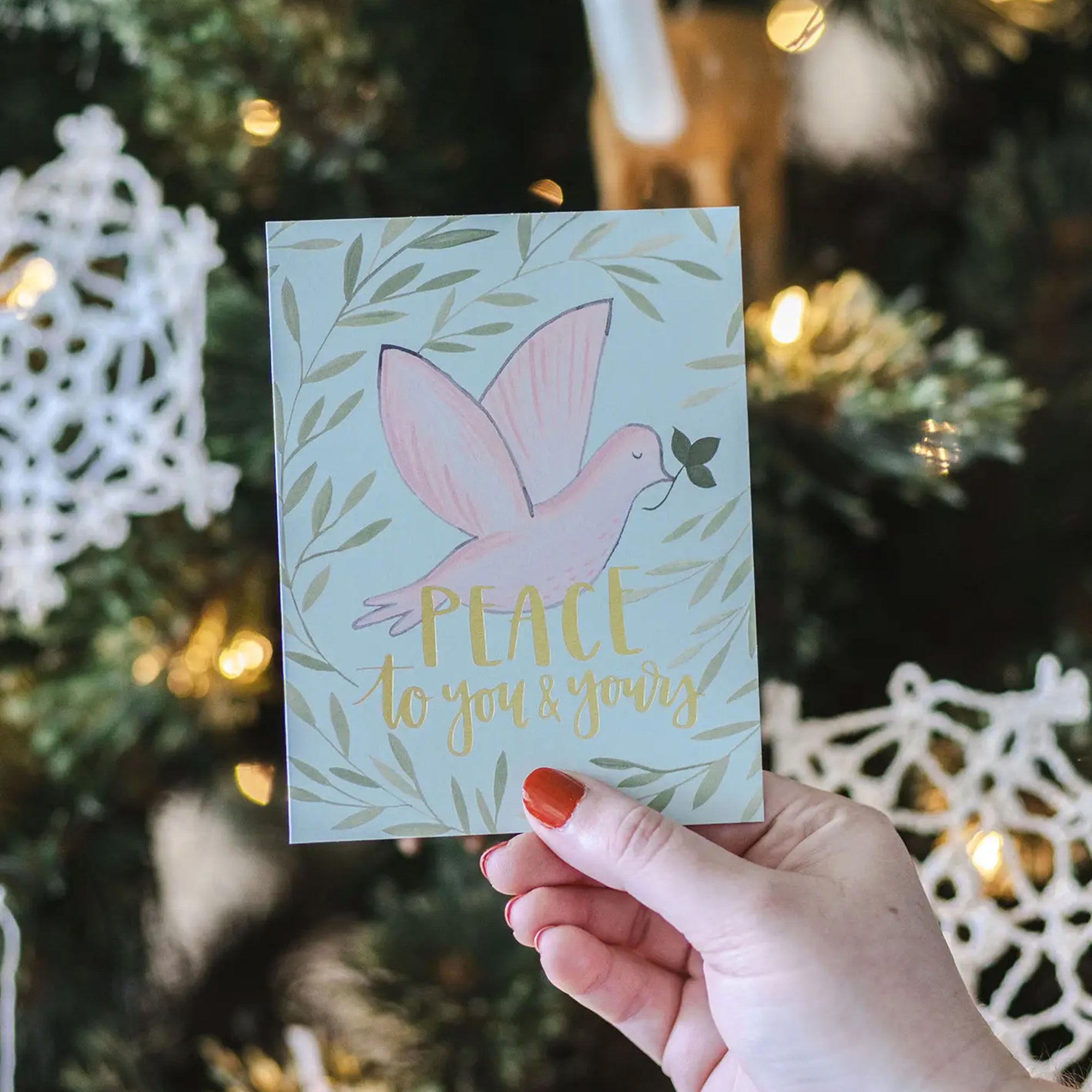 Hand holding a Christmas card with a dove and 'Peace to you and yours' text in front of a decorated tree.