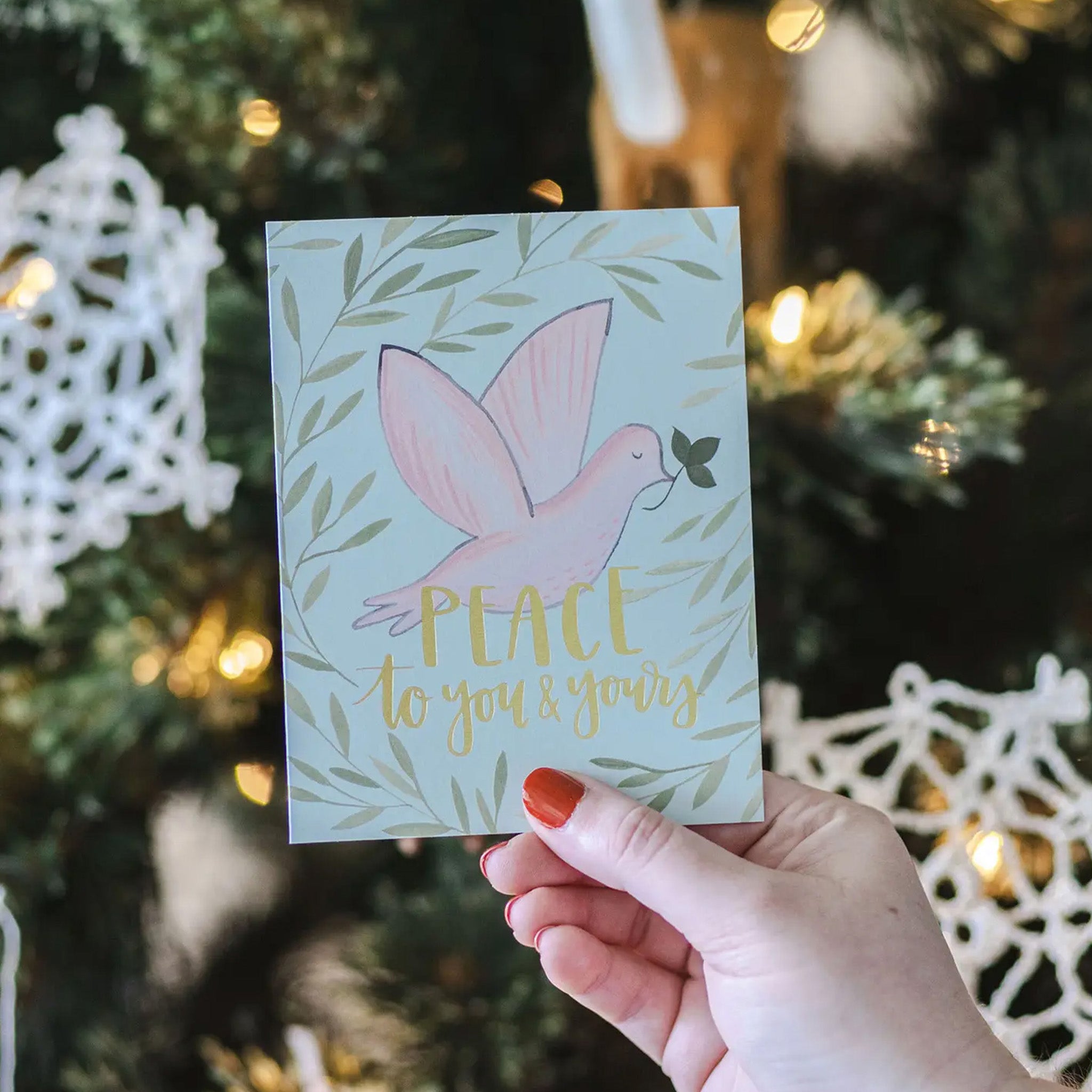 Hand holding a Christmas card with a dove and 'Peace to you and yours' text in front of a decorated tree.