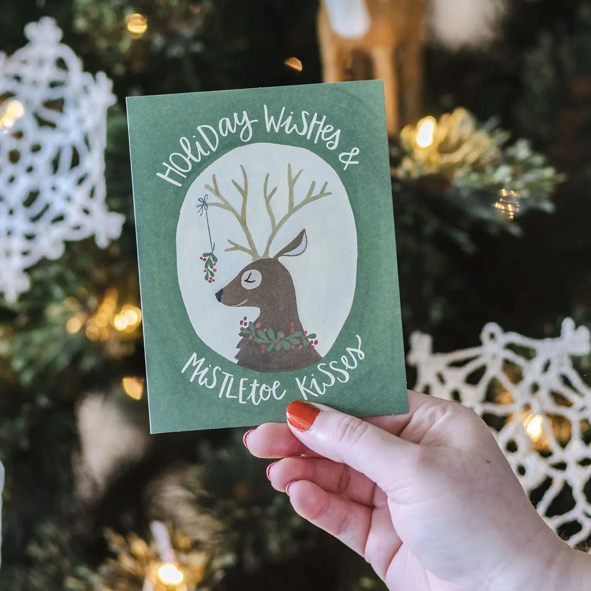Hand holding a green holiday card with a deer illustration in front of a decorated Christmas tree.