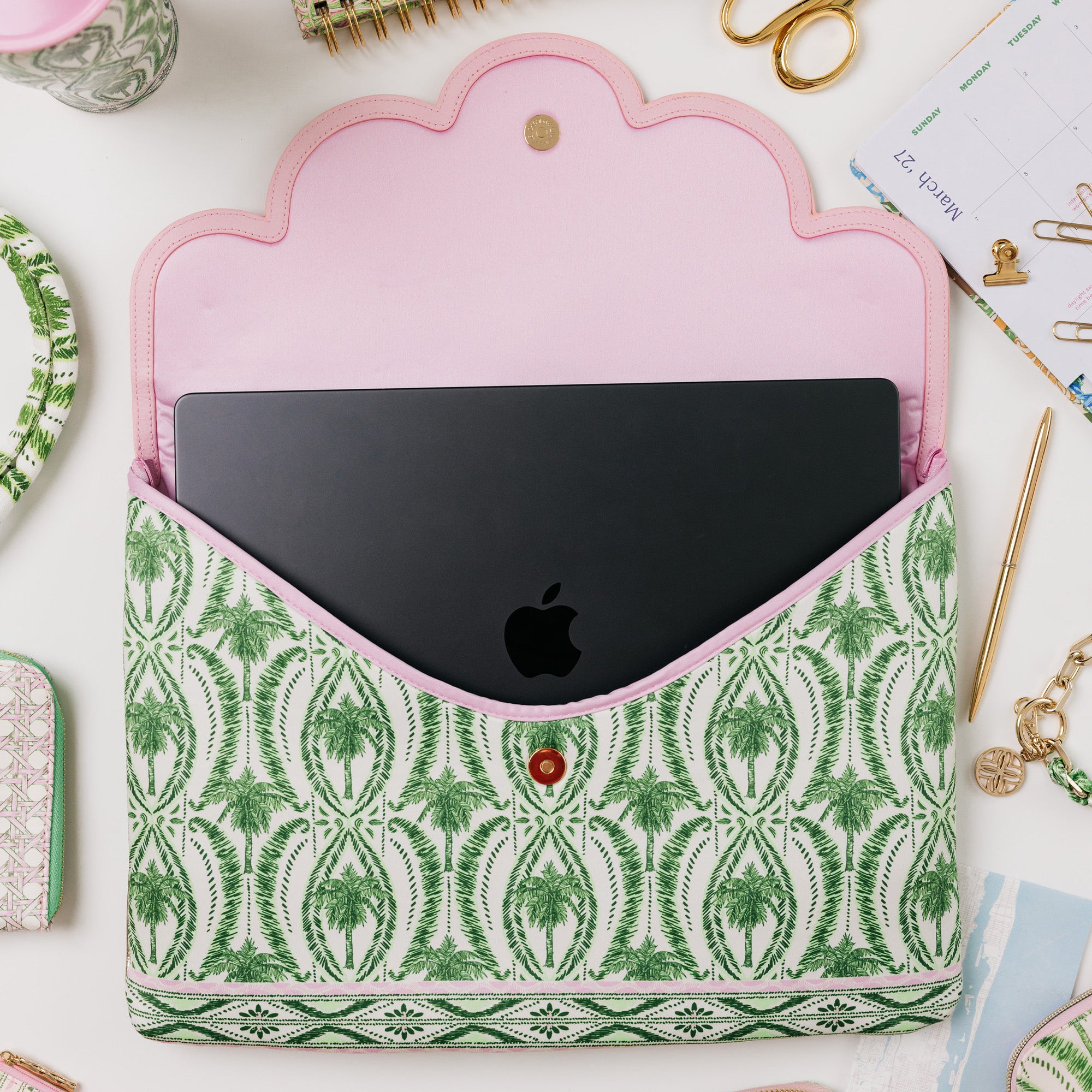 A laptop sleeve with a green and white palm tree pattern and a pink interior, shown holding a black laptop. The sleeve is photographed from above on a white desk with stationery items around it.
