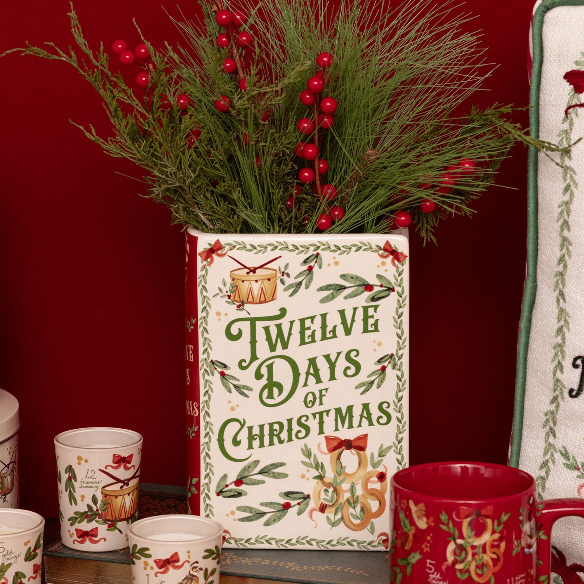 A festive Christmas-themed ceramic tin planter with 'Twelve Days of Christmas' printed on the front, filled with green pine branches and red berries, styled on a shelf with matching red and white mugs against a dark red background.