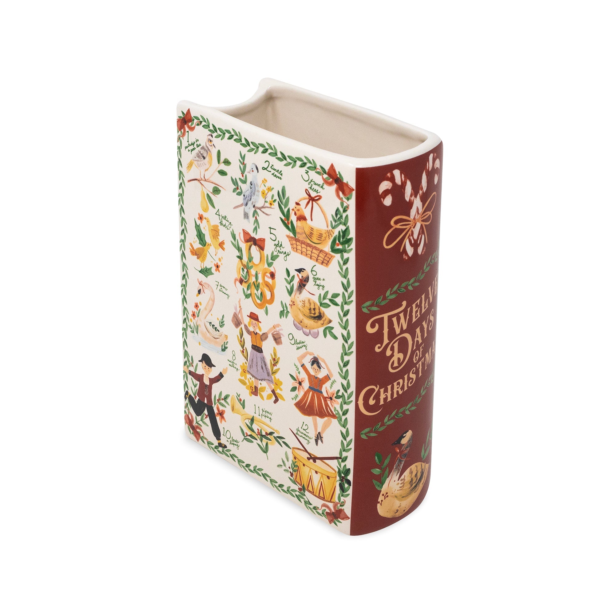 A ceramic Christmas book-shaped vase featuring a cream-colored cover decorated with vintage holiday illustrations and a dark red spine with gold lettering that reads 'Twelve Days of Christmas'.