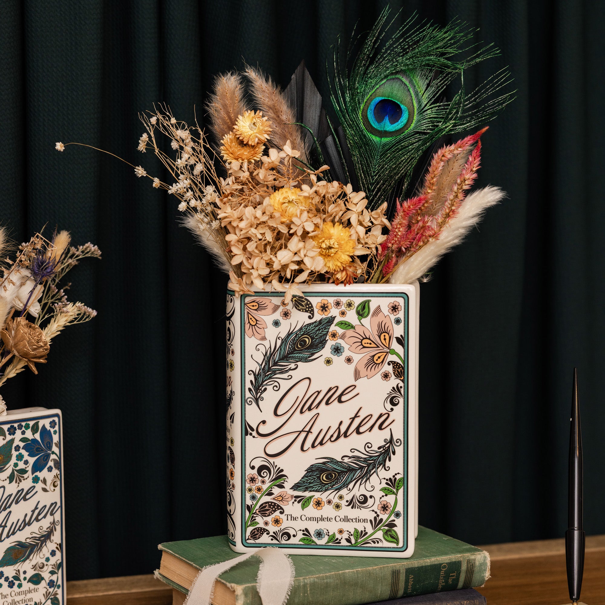 A square ceramic book-shaped vase featuring a white background decorated with pink flowers, green leaves, black swirls, and the text 'Jane Austen' with a feather illustration with dried flowers inside
