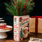 A decorative ceramic Christmas vase with festive red and green holiday illustrations, filled with pine branches and red berries, set on a wooden table with wrapped gifts and holiday decor in the background.