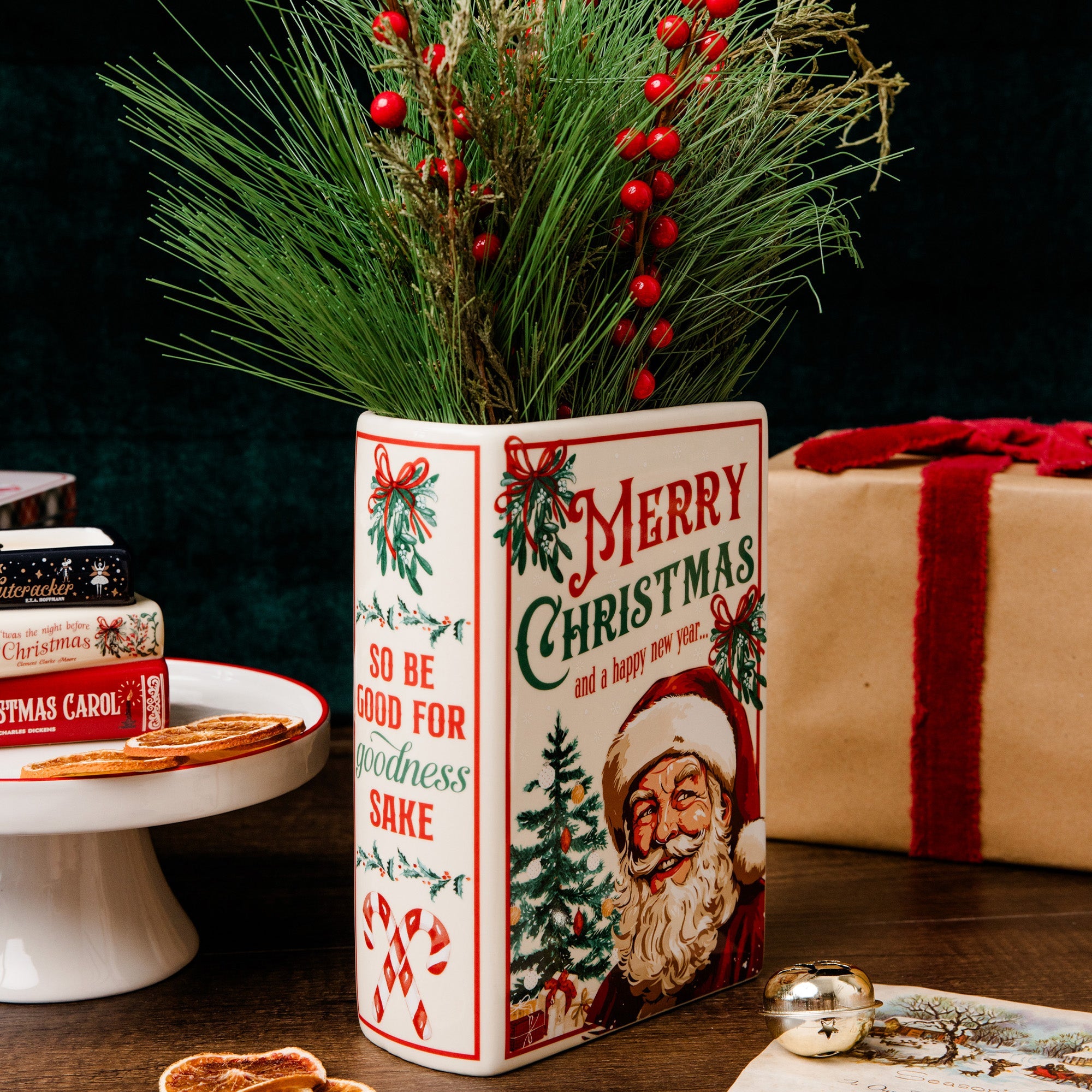 A decorative ceramic Christmas vase with festive red and green holiday illustrations, filled with pine branches and red berries, set on a wooden table with wrapped gifts and holiday decor in the background.