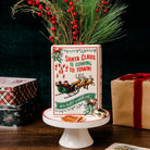 A rectangular ceramic Christmas planter with a festive red and green design reading 'Santa Claus is coming to town!' and 'you better watch out!', displayed on a white pedestal stand with pine branches and red berries placed inside it. Gift boxes and stacked tins sit in the background against a dark backdrop.