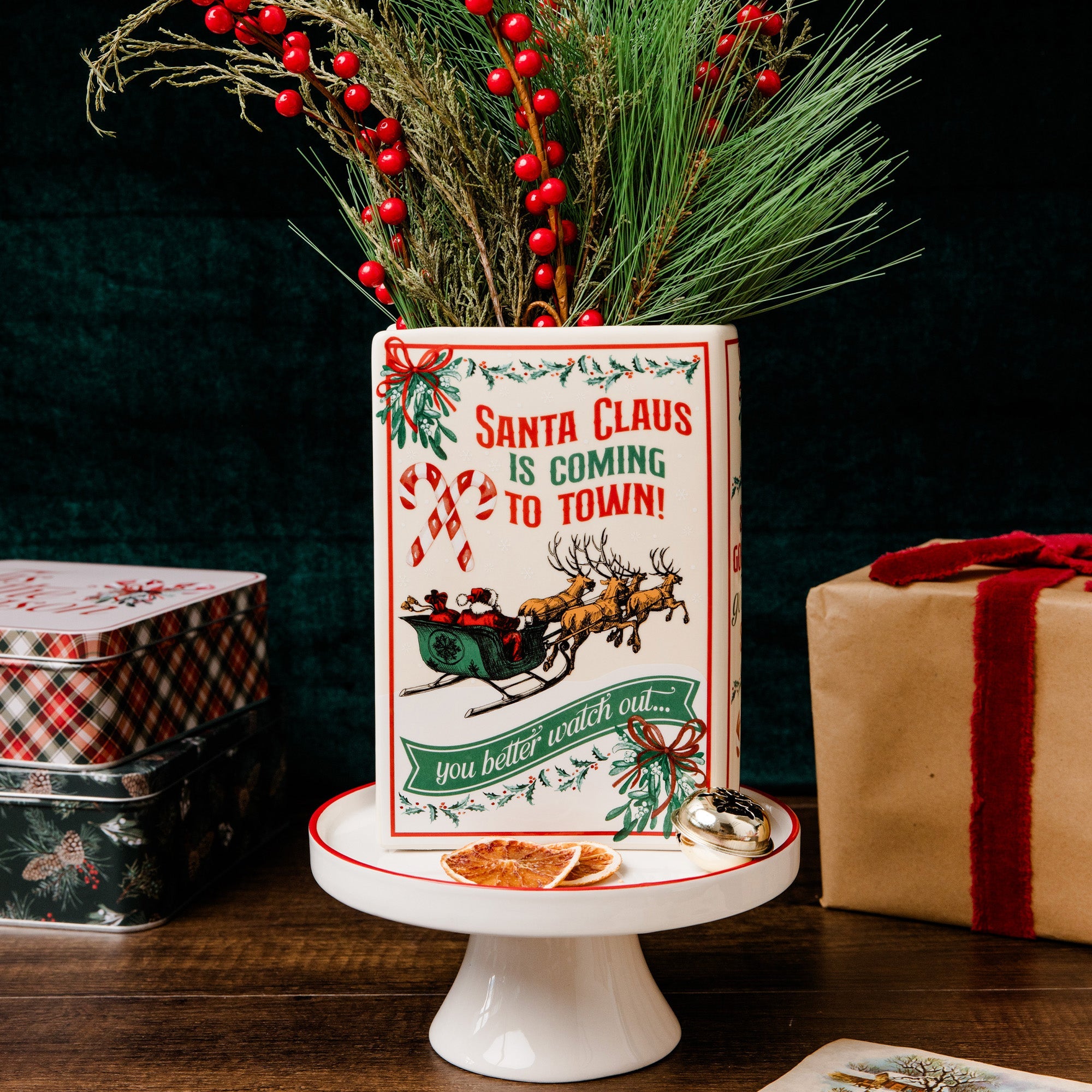 A rectangular ceramic Christmas planter with a festive red and green design reading 'Santa Claus is coming to town!' and 'you better watch out!', displayed on a white pedestal stand with pine branches and red berries placed inside it. Gift boxes and stacked tins sit in the background against a dark backdrop.