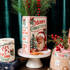 A vintage-style ceramic Christmas canister with a Santa Claus illustration and 'Merry Christmas' text, displayed on a white pedestal stand. The canister is decorated with red berries, pine branches, and holiday greenery, set against a dark green velvet curtain background.