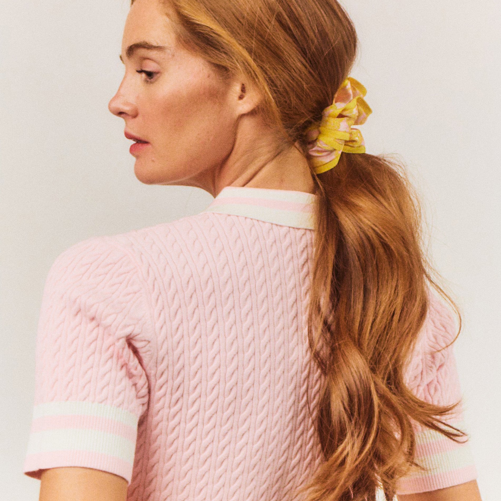 Woman with long hair tied in a ponytail with a yellow scrunchie, wearing a pink cable knit sweater.