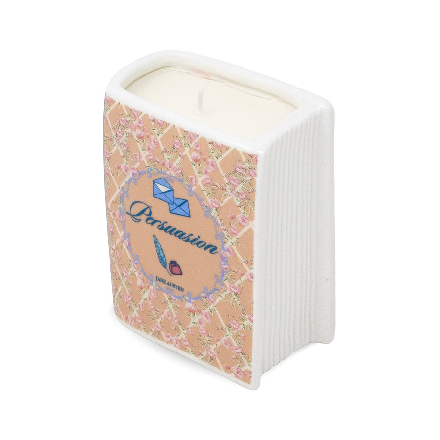 A novelty candle shaped like a book, featuring a white ribbed spine and cover with a pink floral lattice pattern, a central blue circular label reading 'Persuasion', and cream-colored wax visible at the top.