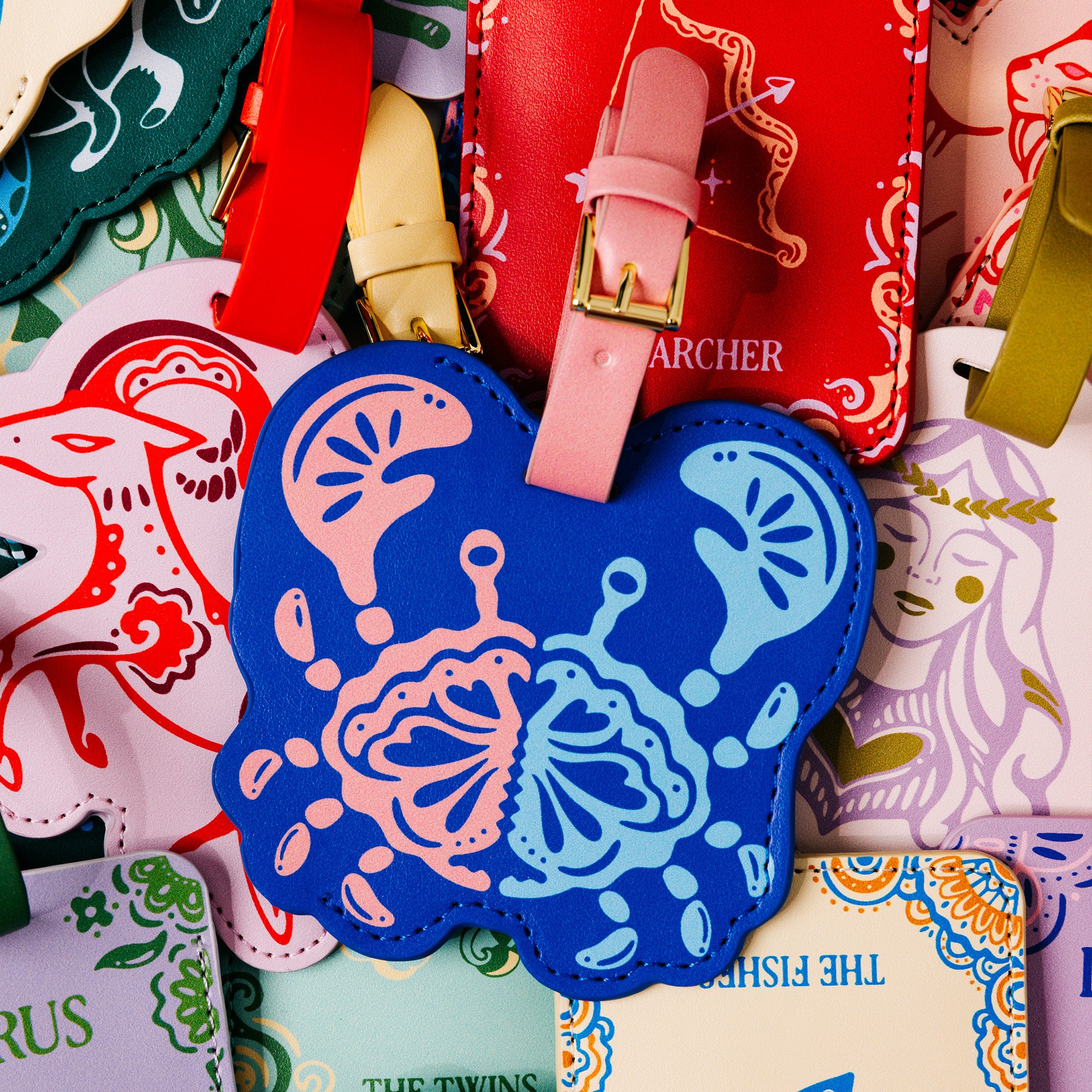 A close-up of a blue cancer crab luggage tag with pink and light blue butterfly designs, layered over other colorful illustrated tags and leather luggage tags with straps.