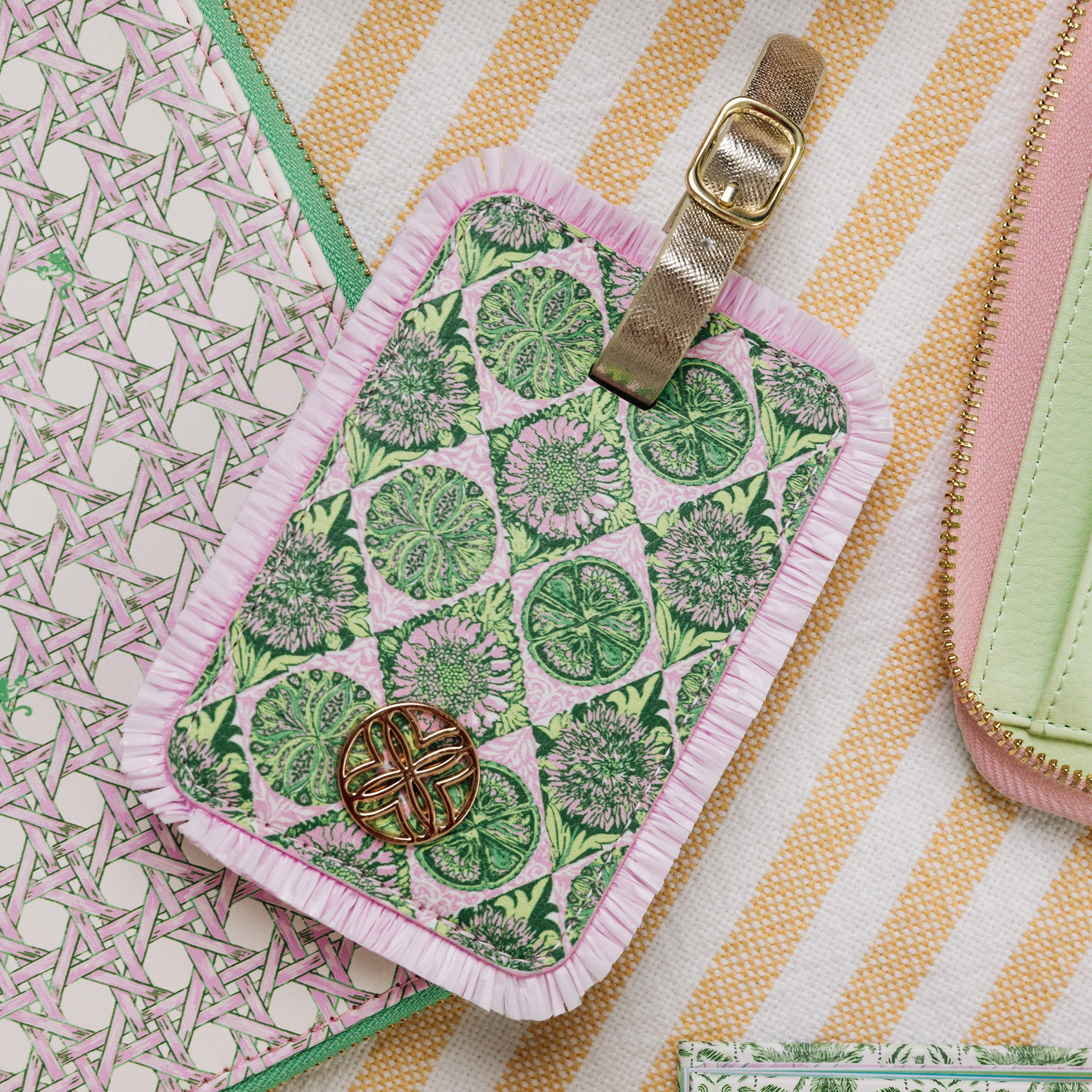 A close-up of a fabric luggage tag with a green citrus and floral print on a pink background, finished with a light pink ruffled border, a beige webbed strap with a gold buckle, and a decorative gold monogram medallion. It is styled on a striped fabric surface alongside other pastel accessories.