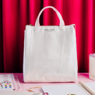 A Kate Spade New York lunch  bag in a textured, vertically pleated off-white fabric, set against a vibrant pink and red curtain backdrop with stationery props on a pink table.